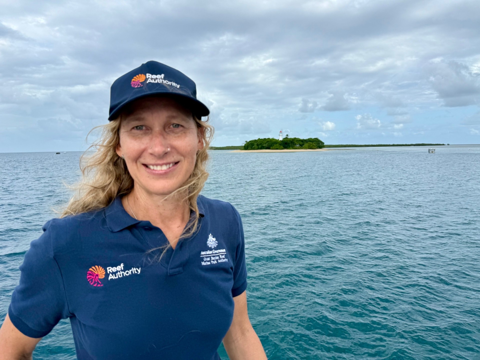 Una mujer con cabello largo y ondulado posa para la cámara con un océano enorme y una pequeña masa de tierra al fondo.