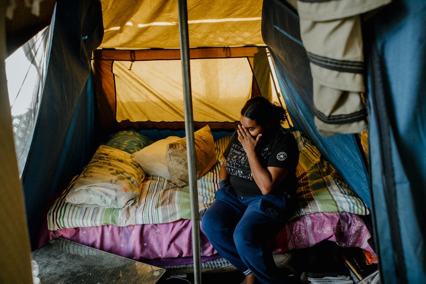 A young woman sits on a bed in a tent, she is putting a hand to her face.