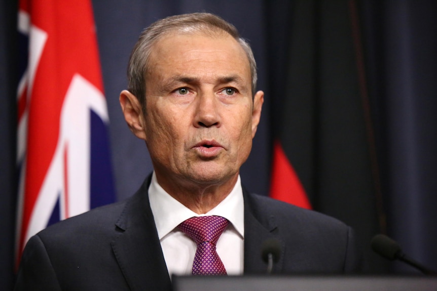 Close-up shot of WA Premier Roger Cook in a suit and tie speaking at a media conference indoors in front of an Australian flag.