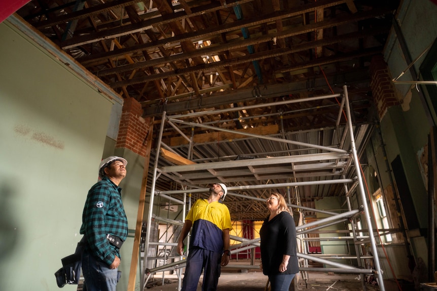 Two men in building clothes and hard hats, one woman survey open ceiling space