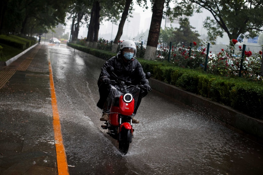 A man wearing a raincoat and face mask rides on a red scooter through a puddle during a storm