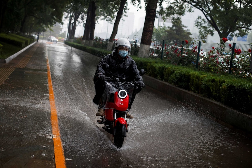 Mężczyzna w płaszczu przeciwdeszczowym i masce na twarz jedzie czerwonym motocyklem przez kałużę podczas burzy