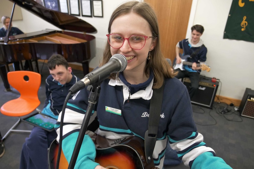 A teenage girl is standing in front of a mircrophone holding a guitar.