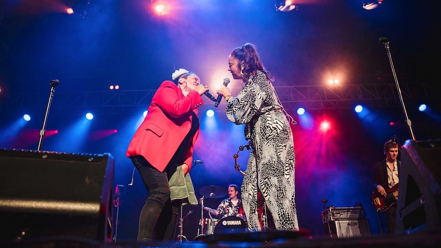 Vika & Linda sing to one another on stage. Vika wears a red jacket, Linda holds a tambourine and wears a jumpsuit.