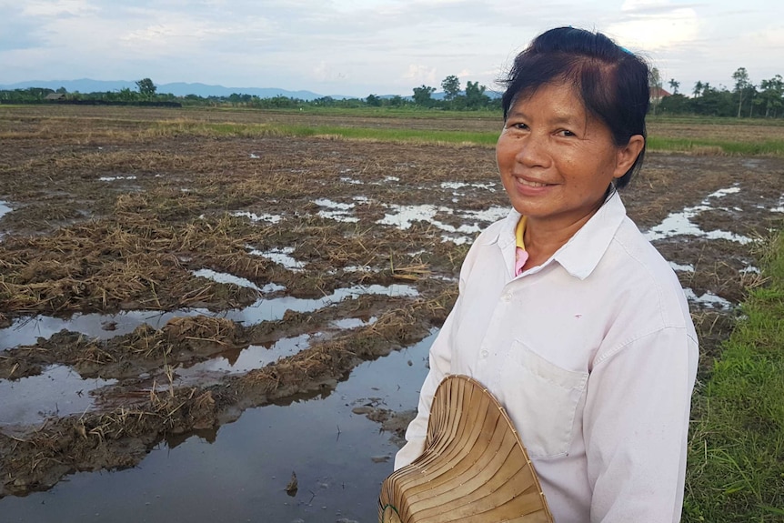 Thai rice farmer Mae Bua Chaicheun stands in a field holding a hat and smiling.