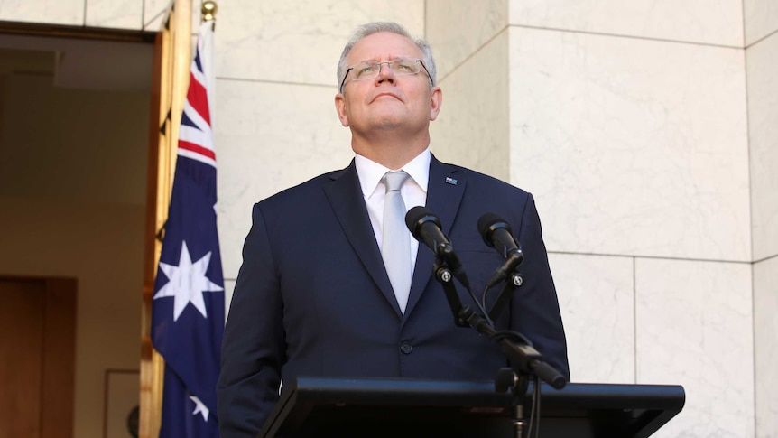 Scott Morrison looks to the sky standing at podium with Australian emblems on them in a courtyard with Australian flags