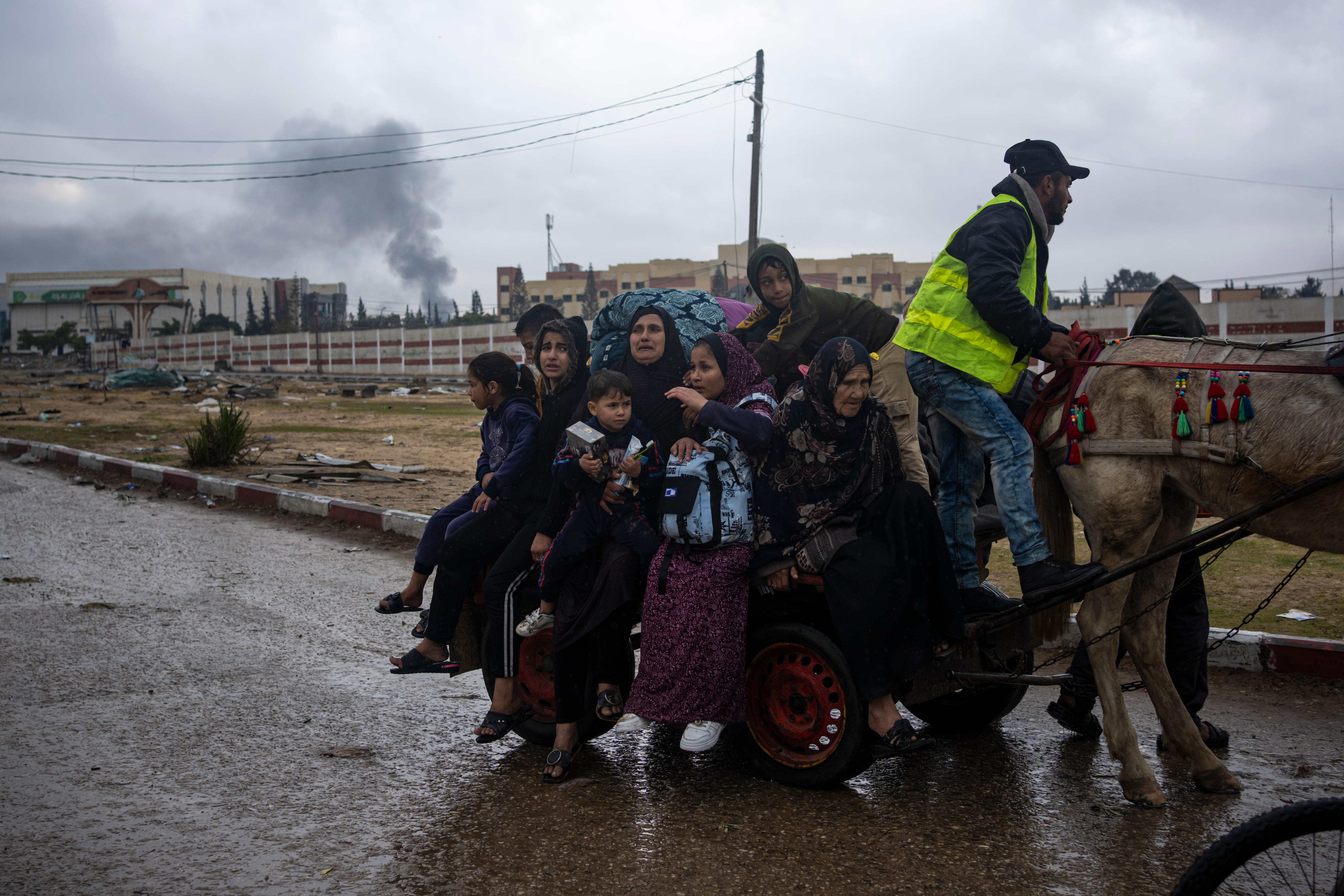 A group of people crowded onto a cart being pulled by a horse. In the background a building smokes. 