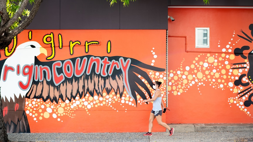 A woman wearing a mask walking past a wall featuring a bright orange mural, in the Darwin CBD.
