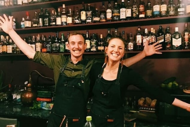 A smiling woman and man behind a bar with bottles of alcohol on the shelves behind them.