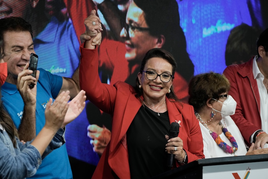 A victorious female political candidate smiles as her running mate holds her right hand aloft as a sign of electoral triumph.