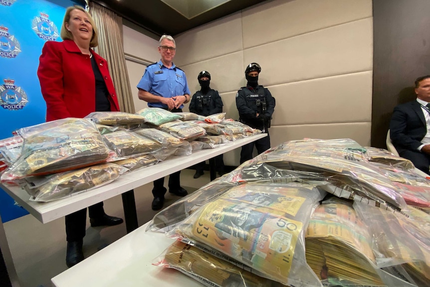 A wide shot of Michelle Roberts with police officers in uniform tactical gear, with piles of money in plastic bags on a table.