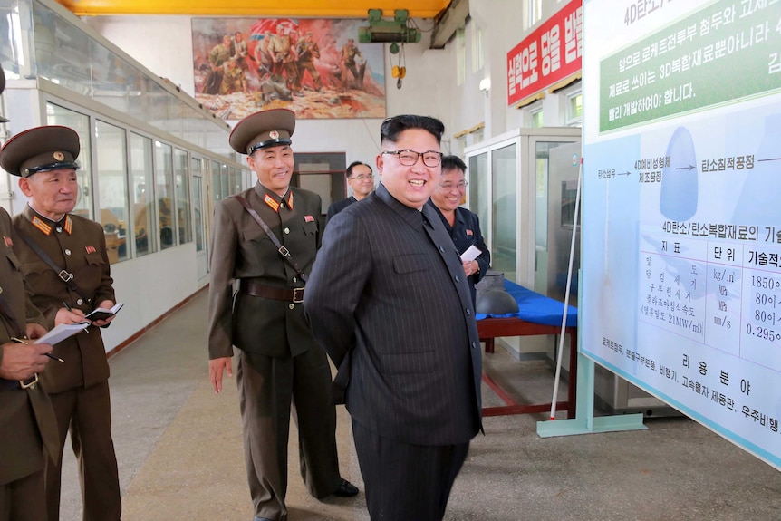 Kim Jong Un smiles and is surrounded by smiling military types as they look at a diagram.