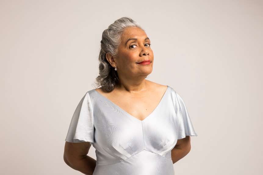 An older Indigenous woman with grey hair in a 50s style, wearing a pale grey 50s-style silk dress.