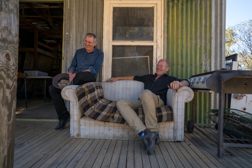 two men sit on the verandah of a ramshackle shed, they are laughing