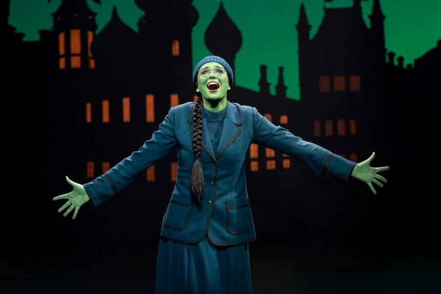 Sheridan Adams holding her arms wide, performing onstage as Elphaba. She is wearing a school uniform and her skin is green.