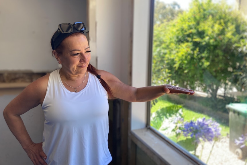 A woman holds her hand out to show a height, next to the window in her house.