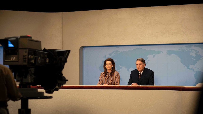 A man and a woman dressed in 1980s clothing sit at a television news desk, looking at a television camera.