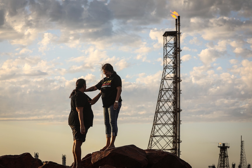 Two women stand with their arms on each other's shoulders, near a gas plant.