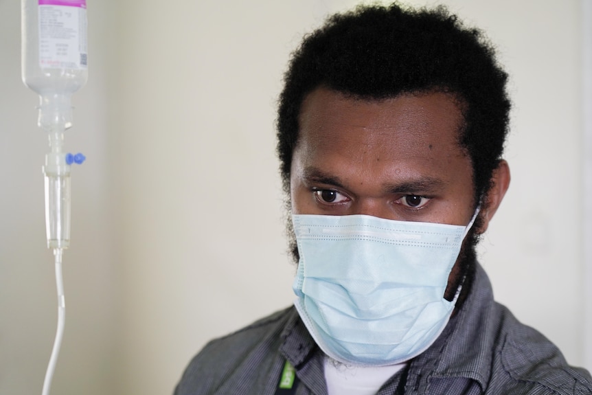 A young man wearing a surgical mask stands next to an IV bag