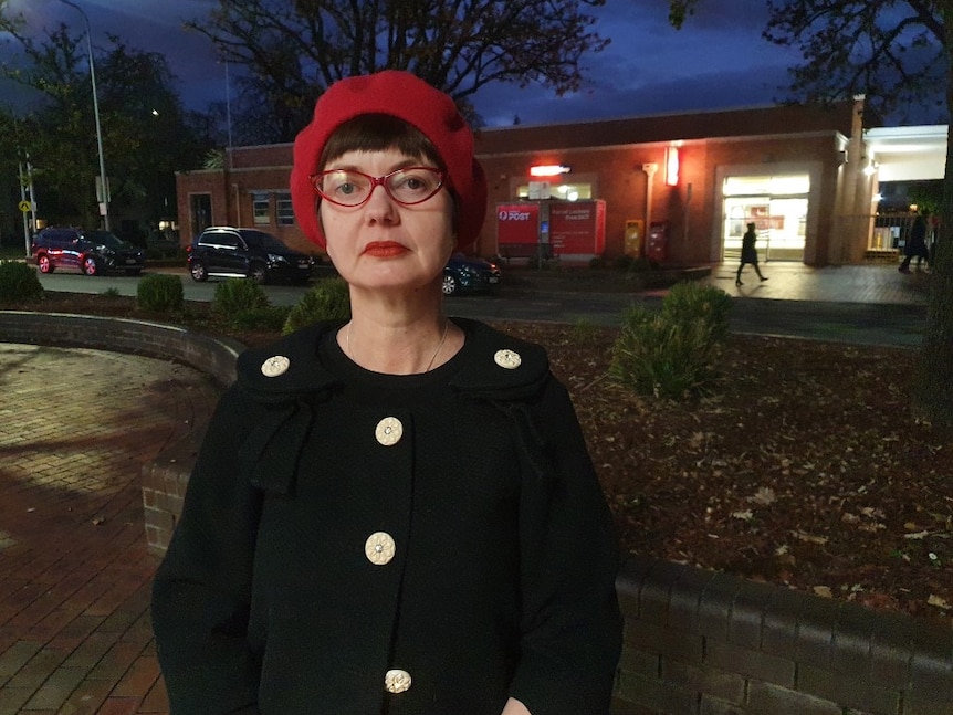 A woman wearing a red beret and red glasses, stands outside the post office at night.
