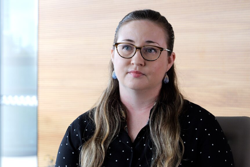 A woman, wearing glasses and a serious face, sits in an office.