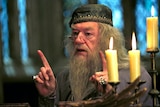 An old man with long hair and beard, wearing a cloth cap, gestures behind a candelabra.