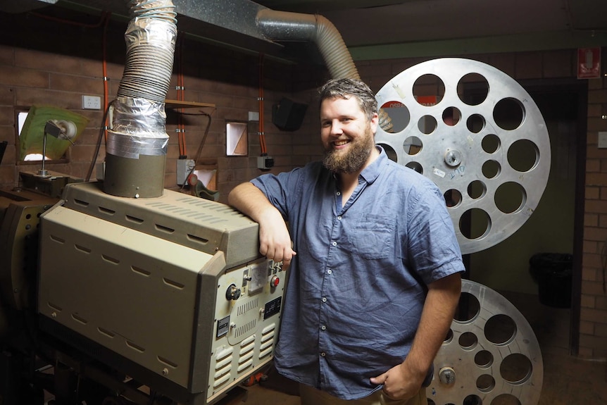 A man in a blue shirt smiles and leans on an old film projector, with two silver film reels behind him.