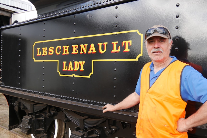 An older man stands next to an old train that has the words Leschenault Lady painted on it.