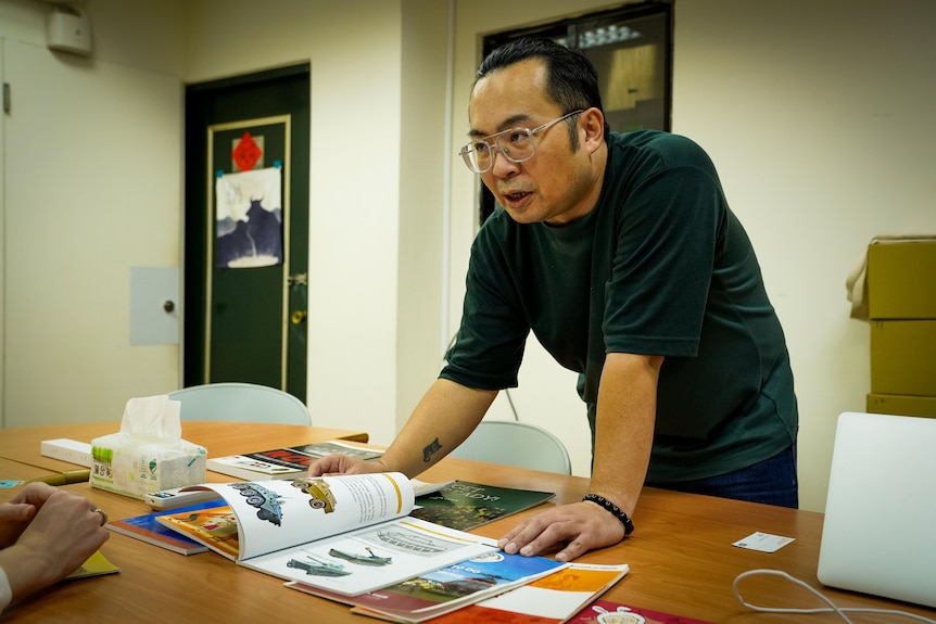 A man wearing green long-sleeved Tshirt leans on a table flipping through pamphlets