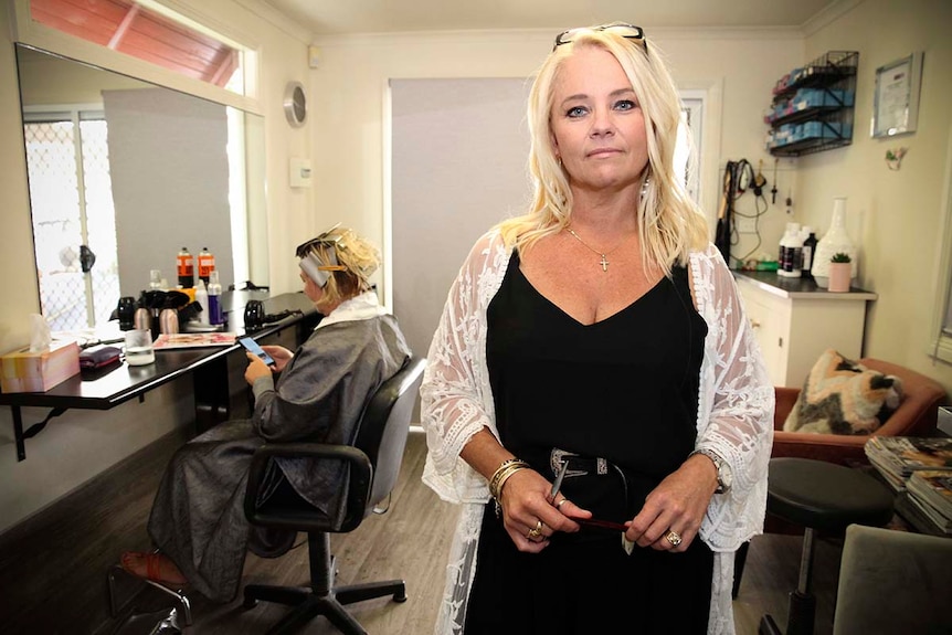 A hairdresser poses in her home salon while a customer sits in her chair in the background
