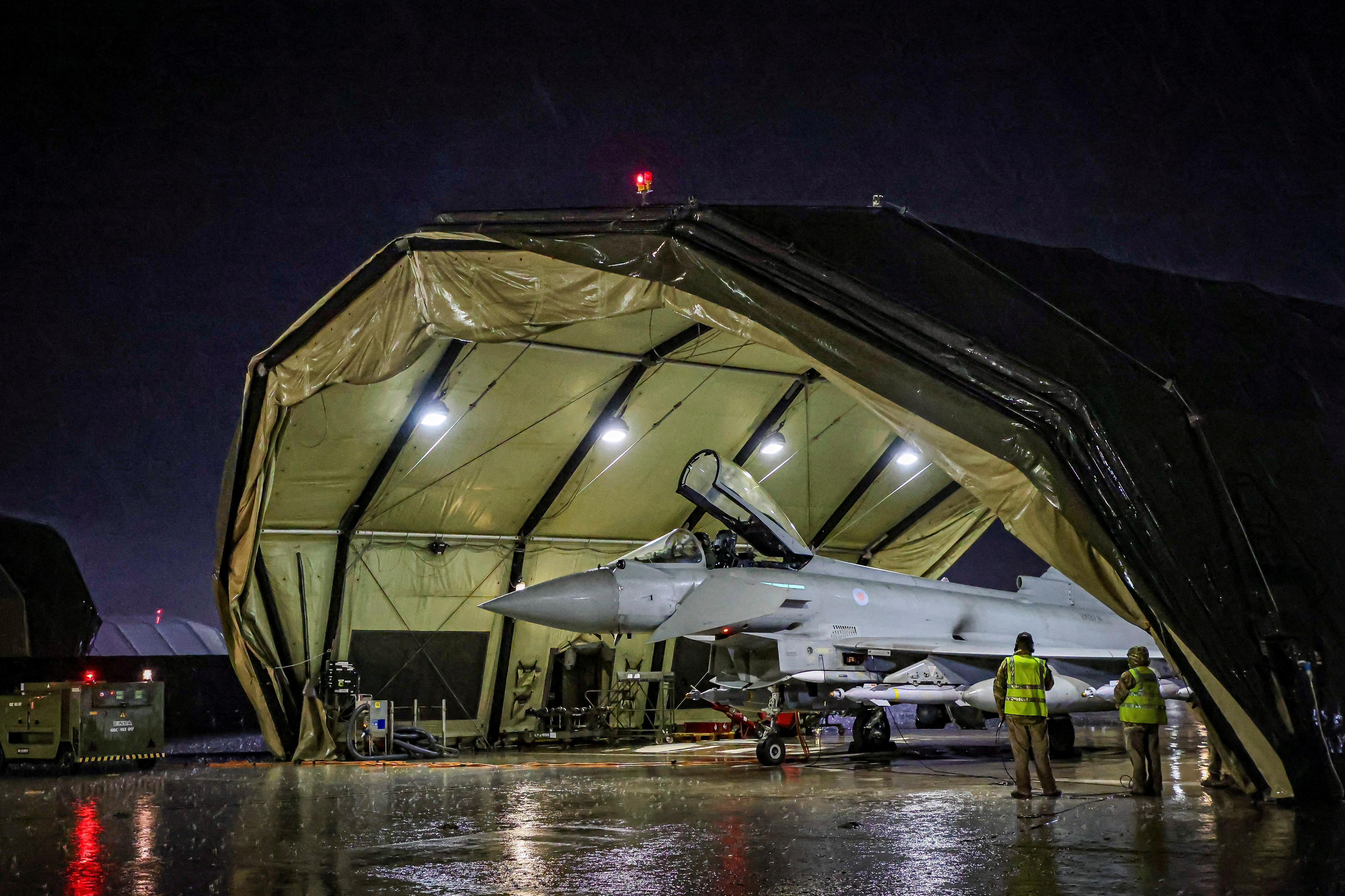  A large grey Royal Air Force aircraft is sheltered by a green canopy with two people in yellow vests standing by it.