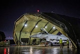  A large grey Royal Air Force aircraft is sheltered by a green canopy with two people in yellow vests standing by it.