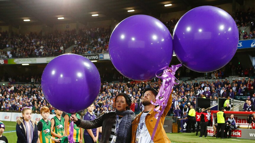 A man and a woman standing in a stadium hold three large purple balloons