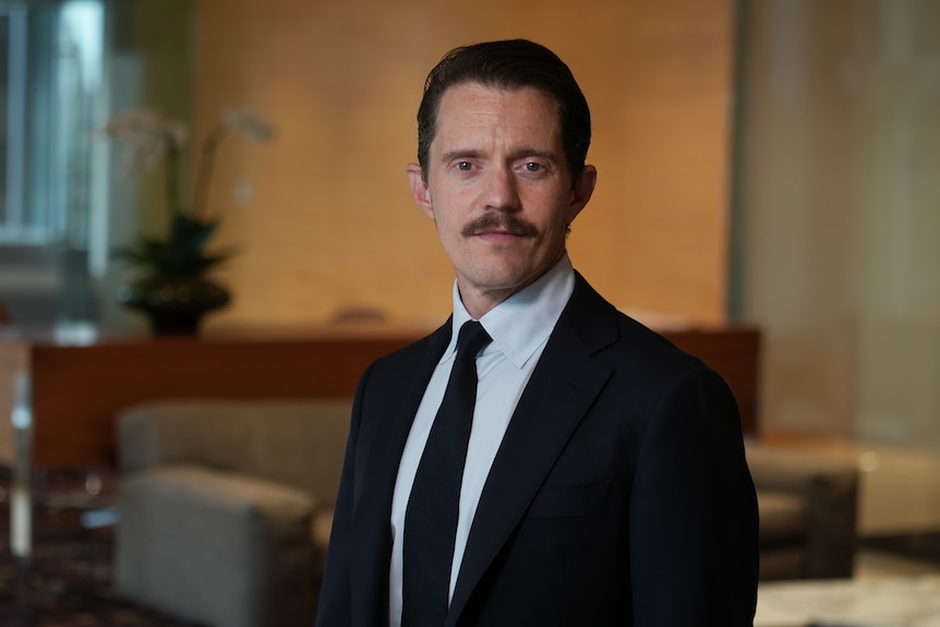 A photo of a young man in a suit, with a moustache, in a board room.