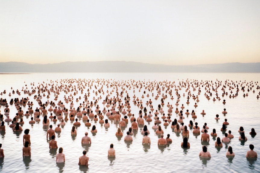 A large number of naked people standing in waist deep water, photographed from behind, looking at hills beyond