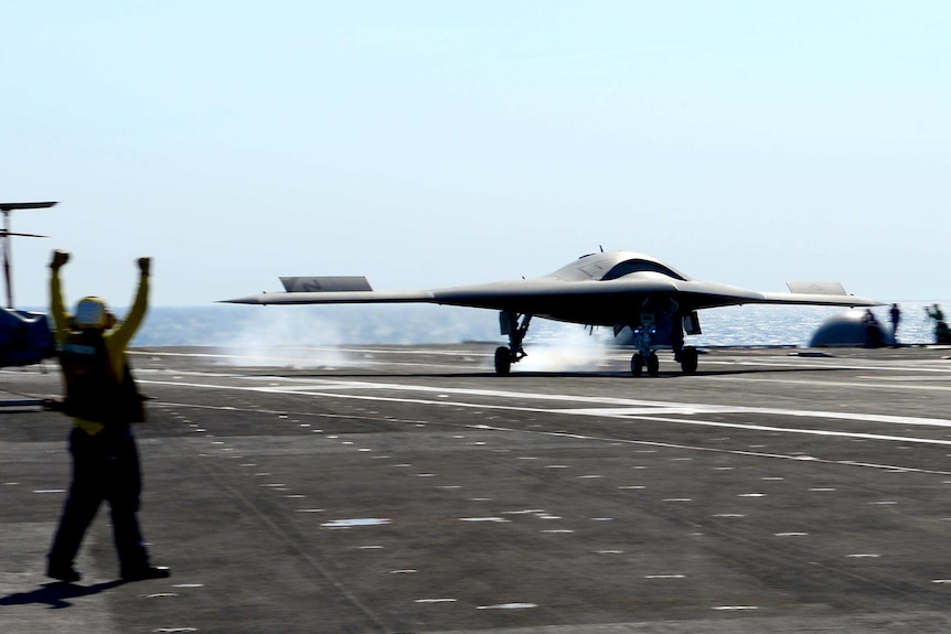 An X-47B UCAS conducts a touch and go landing on the flight deck of an aircraft carrier.