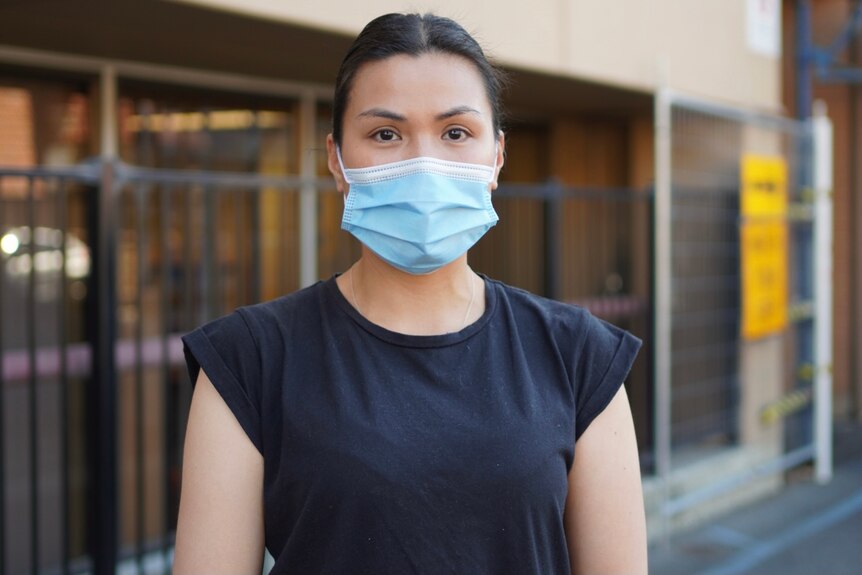 A young woman with black hair pulled back in a bun, wearing face mask and black tshirt.