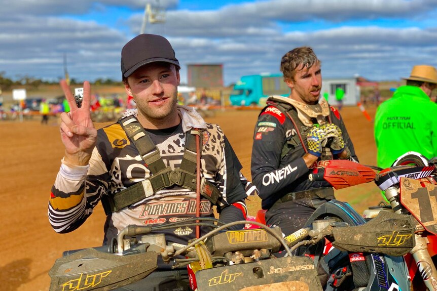 One dirt bike racer sits on his bike in the red dirt, smiles and shows peace sign with another racer in the background