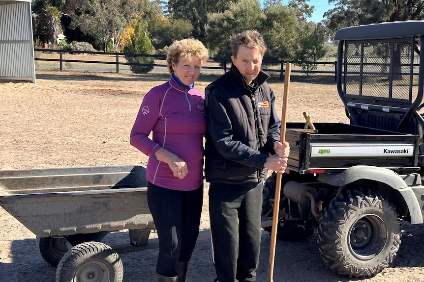 A middle-aged woman and her adult son stand near a small farm vehicle with a trailer attached to it.