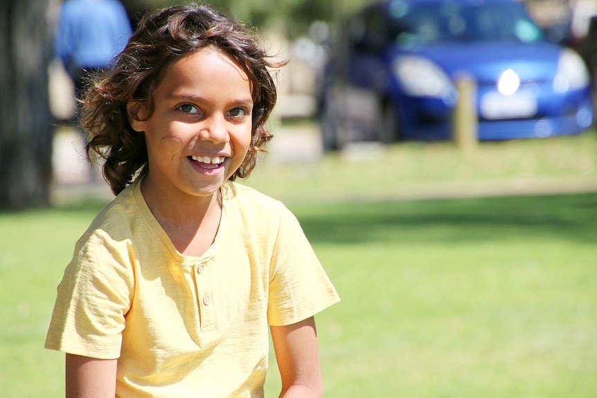 A close up of Tenaya wearing a bright yellow shirt on a grassed oval, smiling.