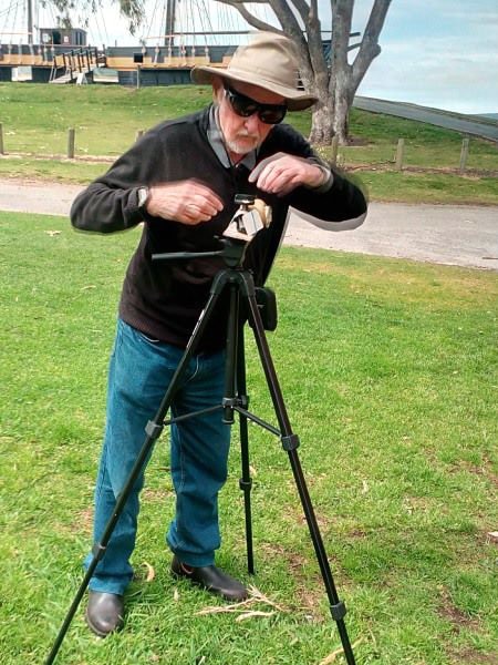 A man standing on grass using a potato camera on a tripod in front of a boat.