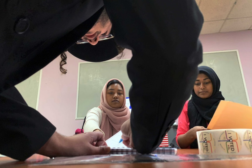 Two women wearing Islamic headwear look on as a man wearing orthodox Jewish clothing holds a pen to paper
