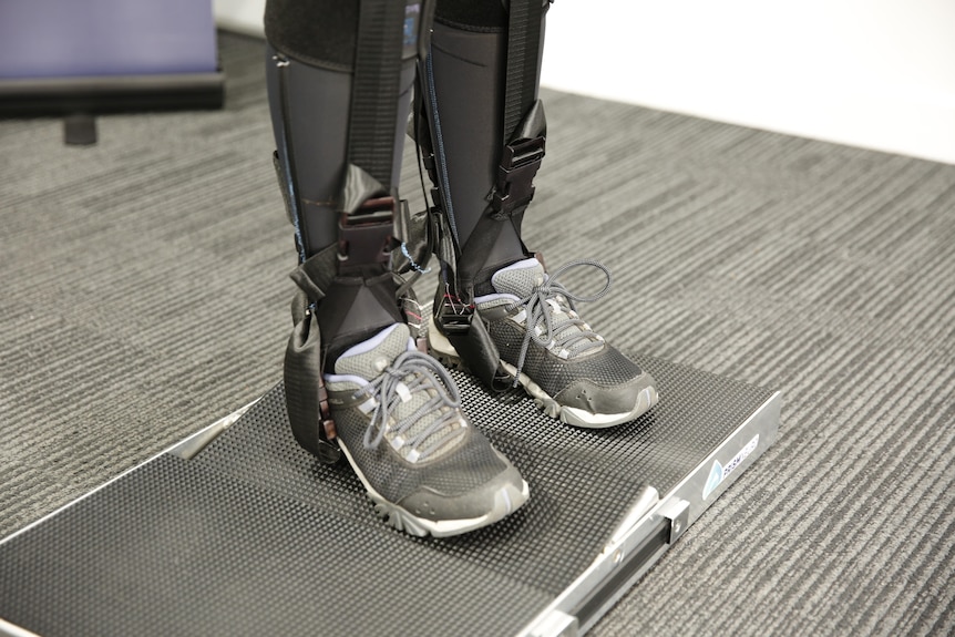 A close up photo of the lower half of a space suit, showing straps and elastic around sports shoes, standing on a scale.