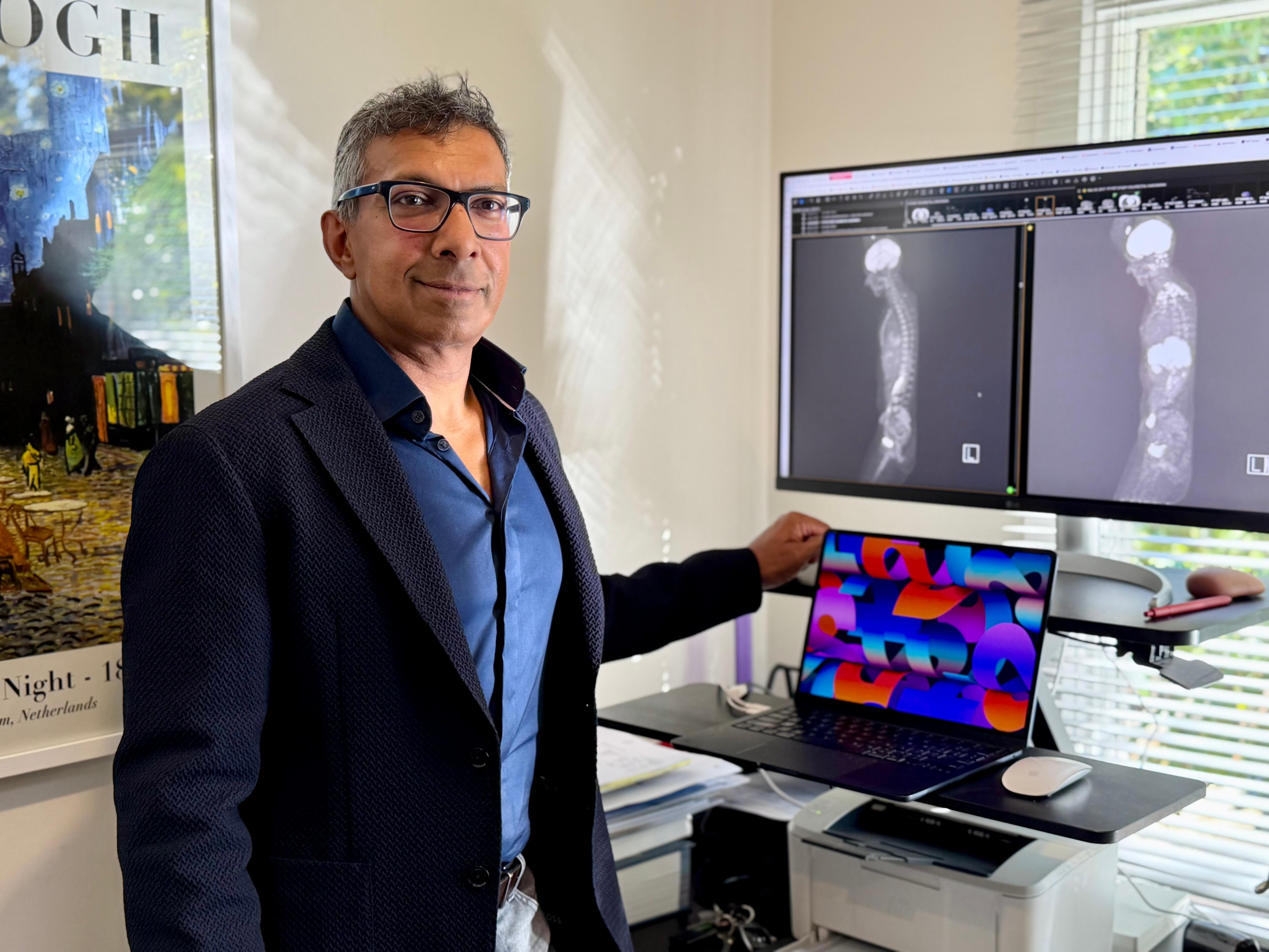 A man in a suit stands in front of a cumputer and some scans of the human body. 