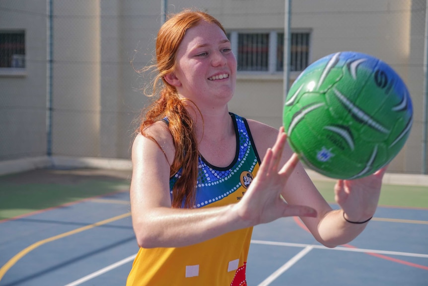 Poppy, with orange-gold hair throws a netball in her colourful Indigenous Budgies uniform
