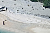 SOS written on a beach, with two people and a boat nearby.