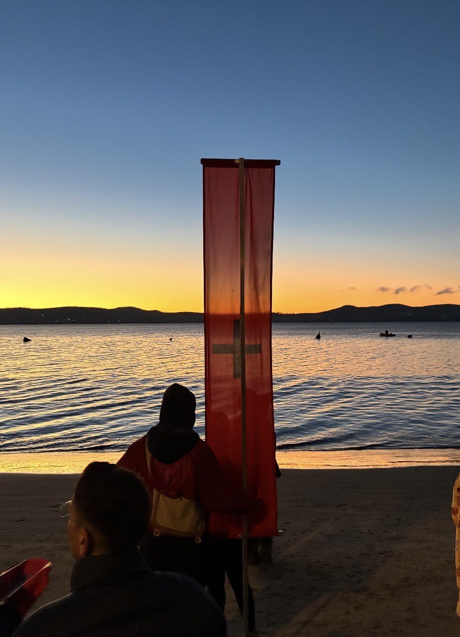 A Dark Mofo red banner with black cross stands on a beach at dawn.