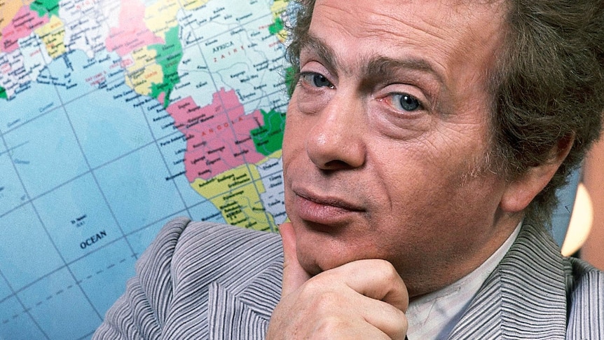 Jackie Mason poses with his hand on his chin and stands in front of a globe.