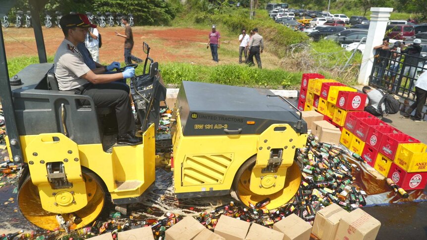Wide shot of a police officer driving a steamroller over rows of glass bottles.