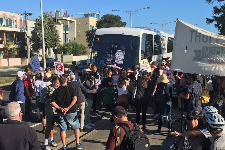 Protesters stand in front of a bus with signs and banners.